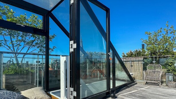 A modern glass-enclosed room with a wooden deck, a wicker chair, and a view of a cityscape. - Heart of the city Penthouse with rooftop terrace (København)