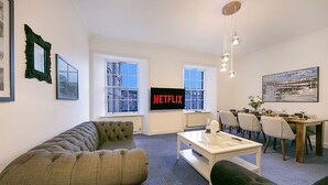 A living room with a sofa, a coffee table, a dining table with chairs, a TV displaying 'NETFLIX', and windows with a city view. - Castle Street Apartments | Grampian Lettings Ltd (Aberdeen)
