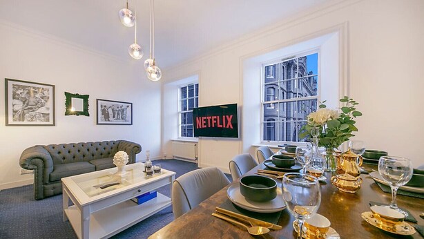 A modern living room with a sofa, coffee table, and dining table set for a meal. A large window offers a view of a cityscape. - Castle Street Apartments | Grampian Lettings Ltd (Aberdeen)