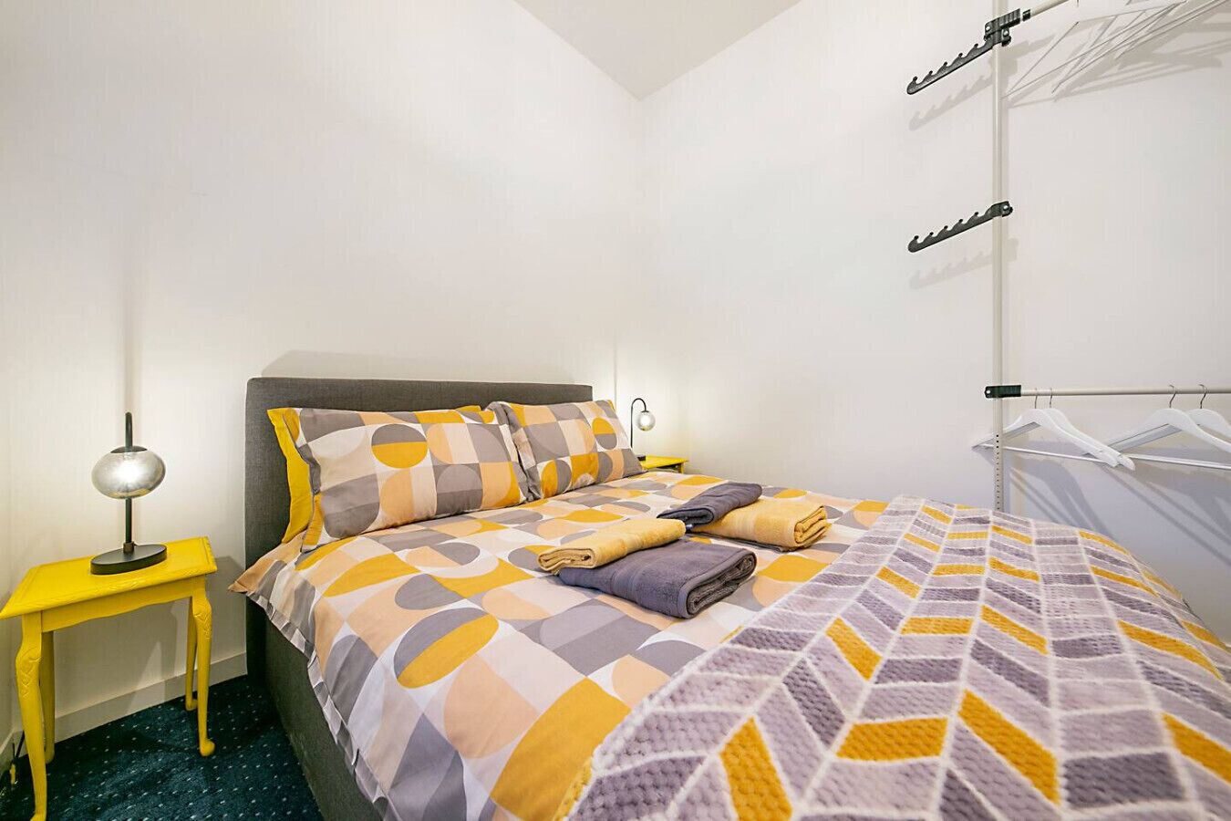A neatly made bed with a patterned bedspread, a bedside table with a lamp, and a wall-mounted drying rack.