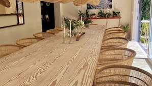 A long wooden dining table with wicker chairs, candles, and plants on a shelf.