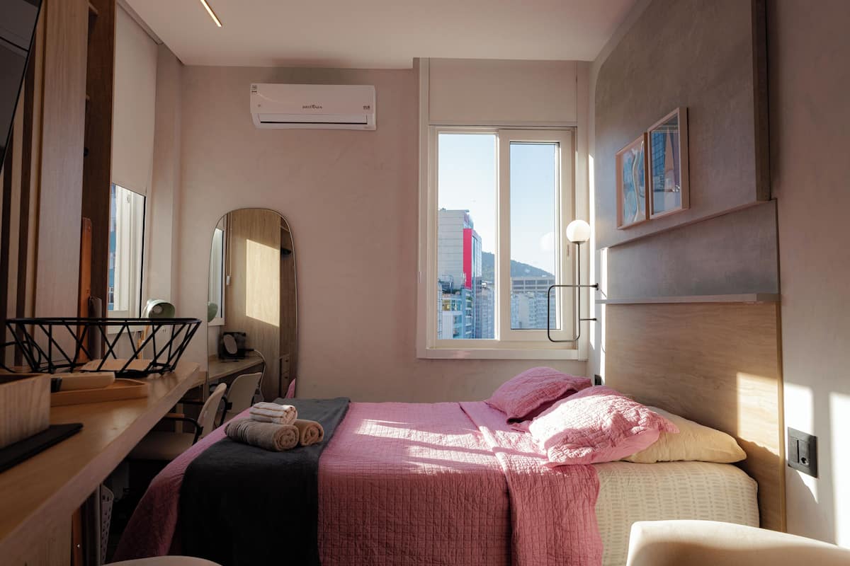Une chambre à coucher comprenant un lit, un bureau, une chaise, un miroir et une fenêtre donnant sur la ville.