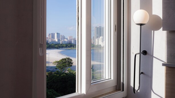 A room with a view of a beach and cityscape through a large window, a modern lamp, and a door with a handle. - 360 Botafogo Belair By SUHCasa (Rio de Janeiro)
