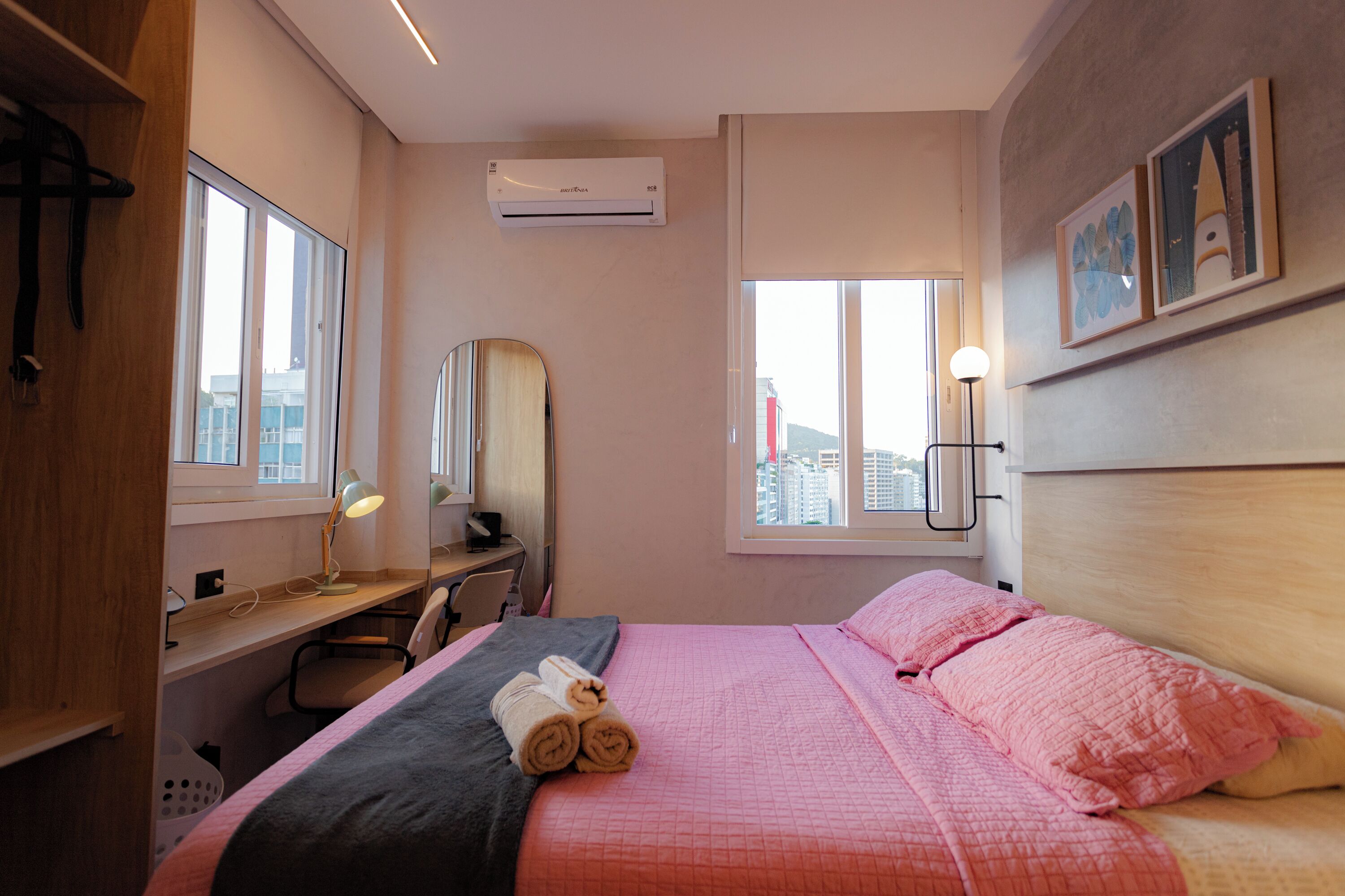 A hotel room with a bed, a desk, a chair, a window with a city view, and an air conditioning unit.