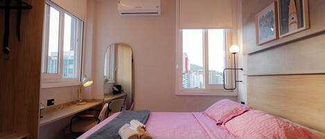 A hotel room with a bed, a desk, a chair, a window with a city view, and an air conditioning unit.
