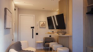 A modern living room with a sofa, two bar stools, a flat-screen TV mounted on a wooden panel, and a small side table. - 360 Botafogo Belair By SUHCasa (Rio de Janeiro)