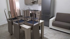 Een moderne keuken met een ingebouwd gasfornuis, een roestvrijstalen koelkast en houten keukenkastjes. Er is een eettafel met vier stoelen en een bank in de woonkamer.