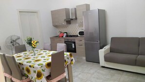 Een kleine keuken met een koelkast, fornuis en magnetron. Er staat een eettafel met een tafelkleed met zonnebloemen en stoelen. Een bank bevindt zich in de woonkamer.