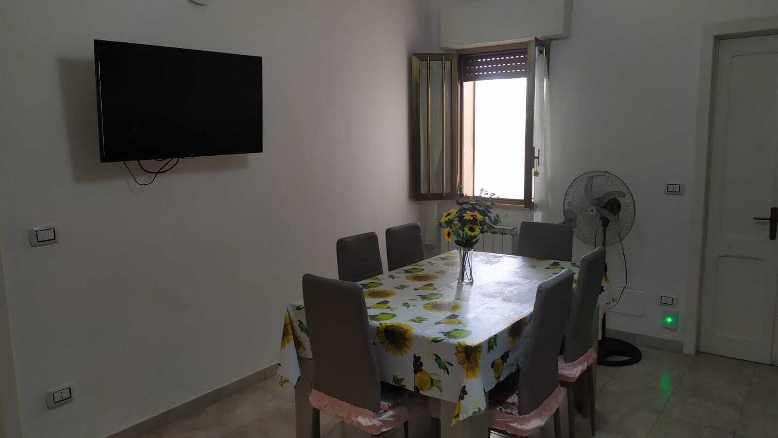A dining area with a table covered in a sunflower tablecloth, surrounded by chairs. A flat-screen TV is mounted on the wall, and a window with curtains is visible.