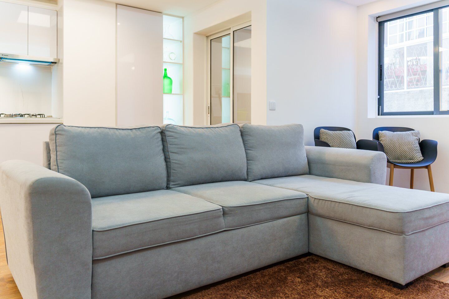 A grey sectional sofa in a modern living room with a brown carpet, a window with a view, and a door leading to another room.