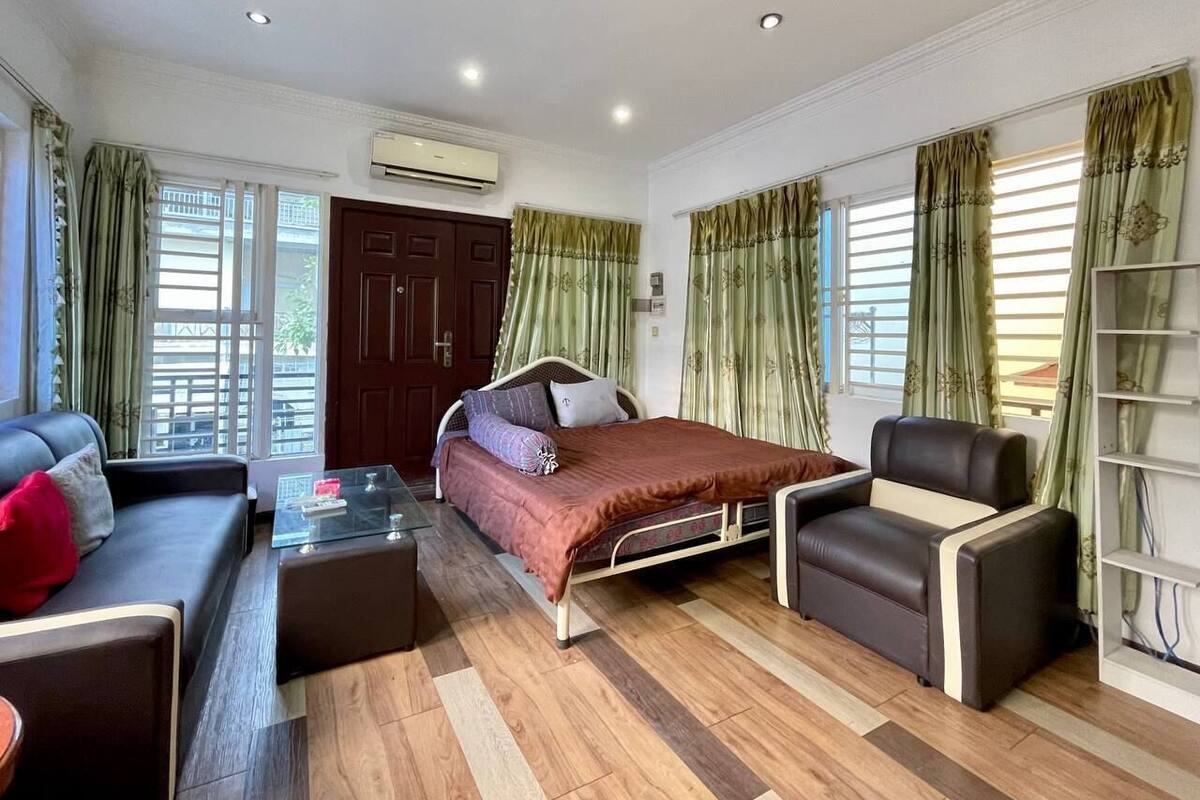 A room with a bed, a sofa, a chair, and a small table. There is a window with curtains and an air conditioning unit on the wall.