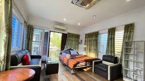 A well-lit room with a bed, sofa, and armchair, featuring a wooden floor and large windows with curtains. - Cassiarata (Phnom Penh)