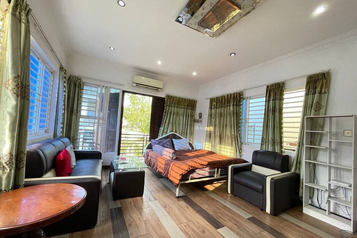 A well-lit room with a bed, sofa, and armchair, featuring a wooden floor and large windows with curtains.
