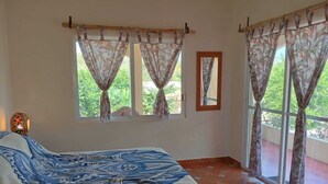 A bedroom with a bed, two windows with curtains, a mirror, and a tiled floor.