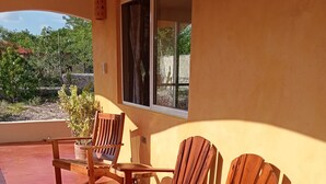 A patio with wooden chairs and a table, a window with curtains, and a view of greenery outside.
