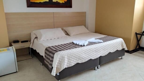 A neatly made bed with a quilted cover, a bedside table with a lamp, and a painting of a sunset on the wall. - HOTEL SAN JERONIMO POPAYAN (Popayán)