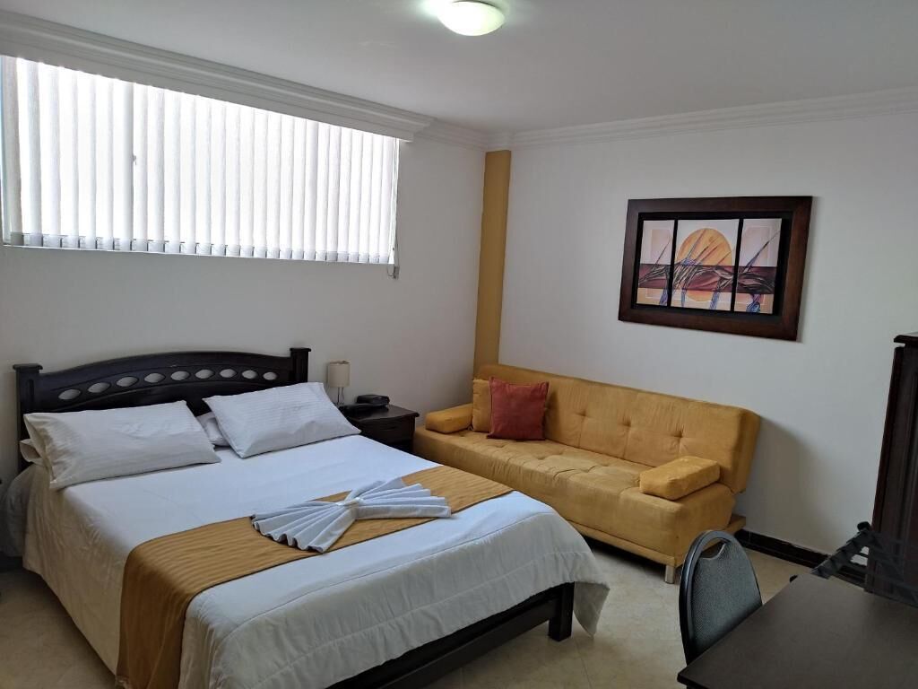 A hotel room with a bed, a sofa, a painting on the wall, and a window with blinds.