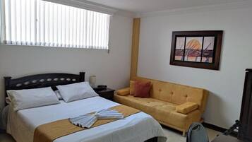 A hotel room with a bed, a sofa, a painting on the wall, and a window with blinds.