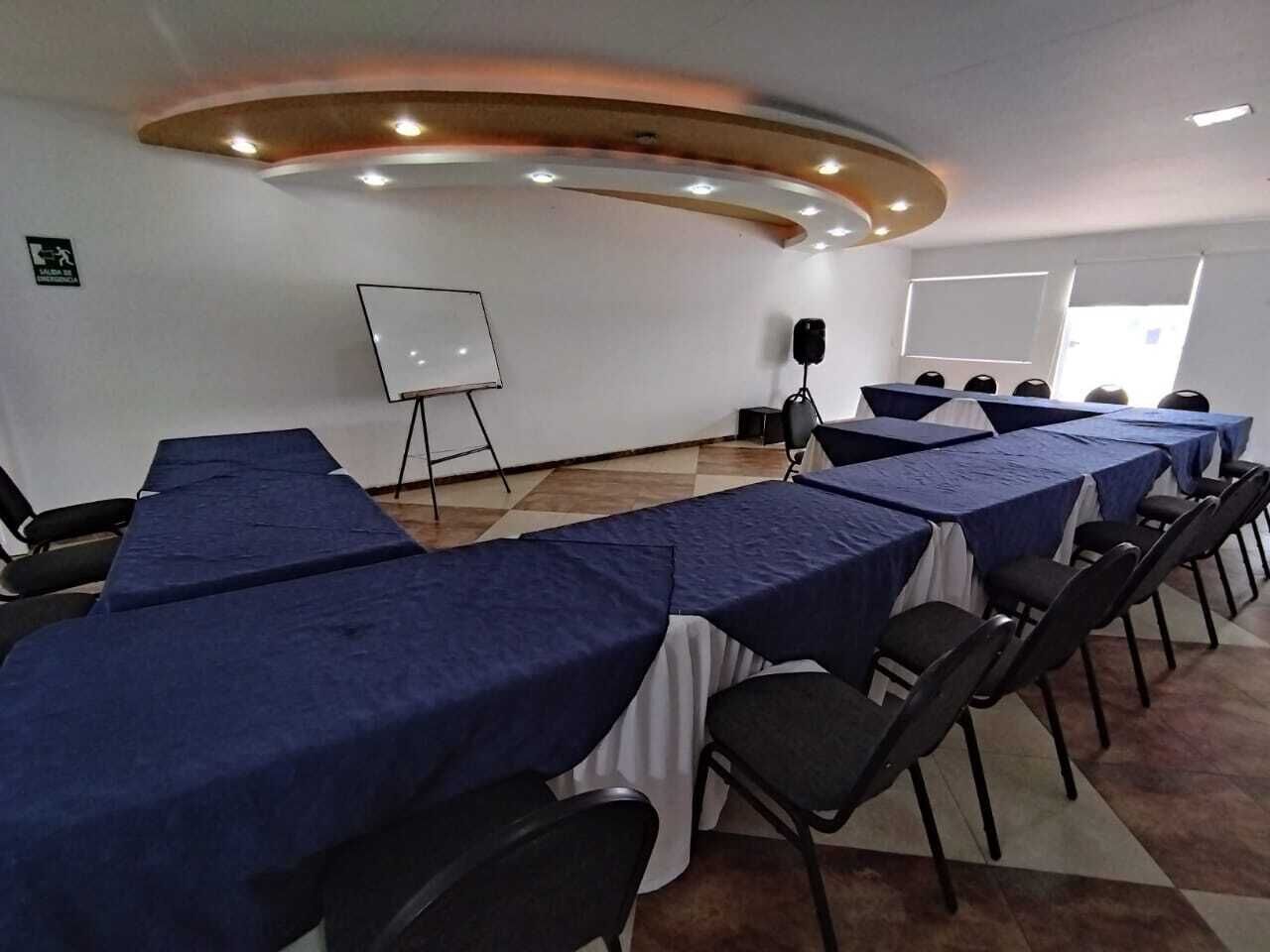 Un salón de conferencias con largas mesas cubiertas con manteles azules, sillas negras, una pizarra blanca y un altavoz sobre un atril.