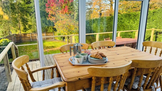 En træterrasse med et bord dækket til morgenmad, blandt andet croissanter og kopper, med udsigt over en have med træer og en sø.