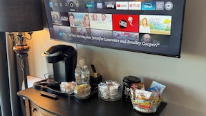 A hotel room with a flat-screen TV displaying a streaming service interface, a coffee maker, a remote control, and a snack basket on a wooden dresser.