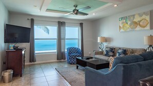 A living room with a television, a sofa, a coffee table, and a chair. There are curtains on the windows, a ceiling fan, and a painting on the wall. - 3-bedroom, 3 bath condo in beautiful Longboat Key. Beachfront views w/ pool. (Longboat Key)