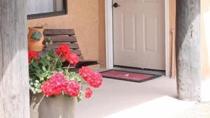 Une maison avec une porte blanche, un banc en bois et un jardinier avec des fleurs rouges.