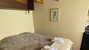 A bedroom with a large bed, a wooden ceiling, a small framed picture on the wall, and a kitchen area visible through an open door.