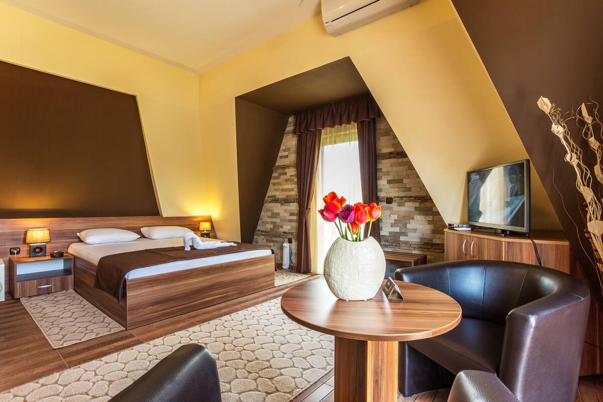 A modern hotel room with a large bed, a sitting area with a table, a TV, and a decorative vase with flowers.