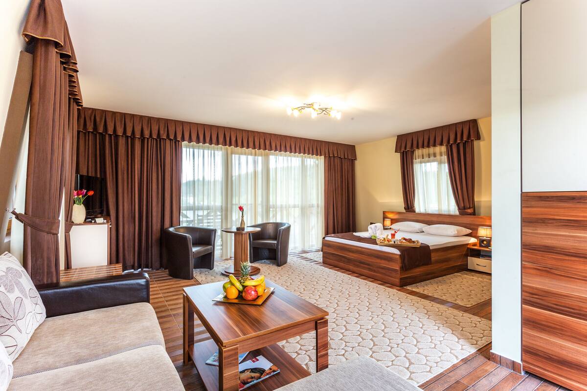 A hotel room with a bed, a sofa, a dining table with fruit, a small table with a vase, and a mirror.