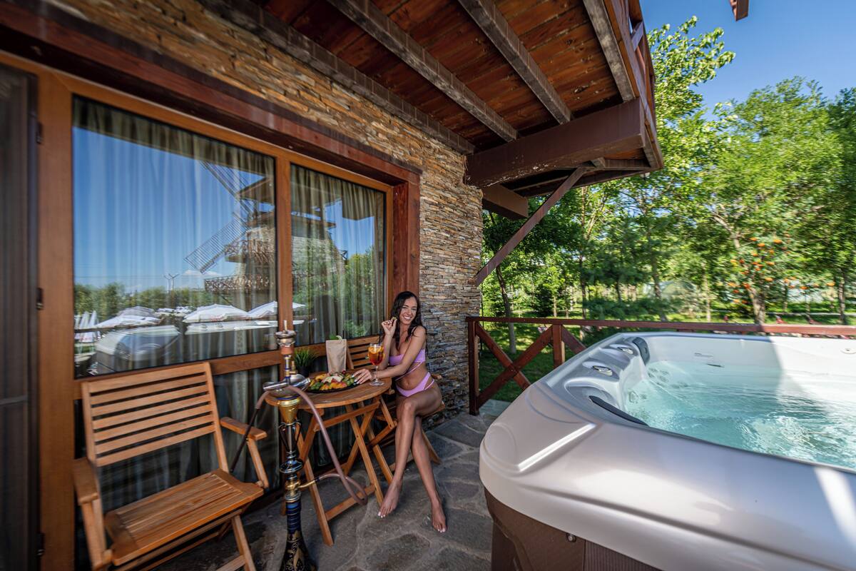 A woman sitting at a wooden table with a hot tub and a wooden chair in a stone-walled room with large windows.