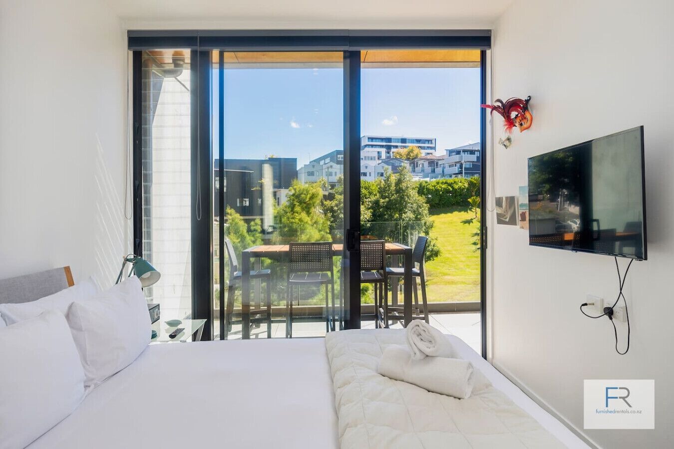 A hotel room with a bed, a TV, a balcony with a table and chairs, and a view of buildings and greenery.