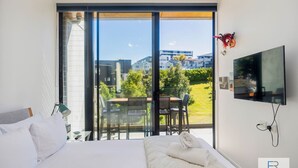 A hotel room with a bed, a TV, a balcony with a table and chairs, and a view of buildings and greenery.