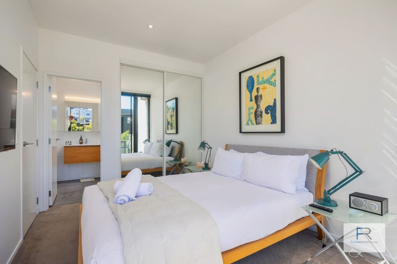 A modern hotel room with a large bed, a desk with a lamp, a mirror, and artwork on the wall.