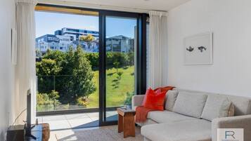 A modern living room with a large sliding glass door leading to a balcony, a grey sofa with red cushions, and a small wooden side table.