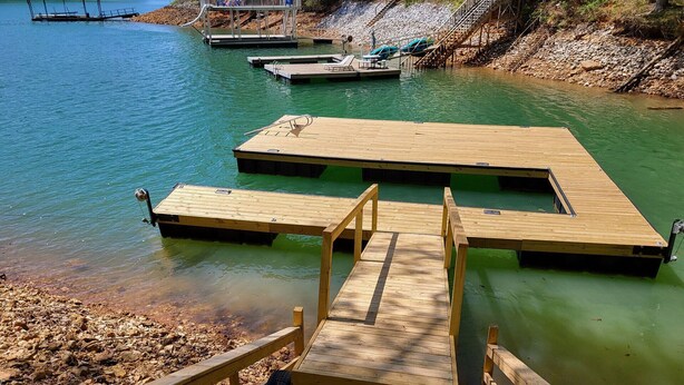 Une jetée en bois s’étendant sur une étendue d’eau calme, avec une rive rocheuse et un petit pont en arrière-plan.