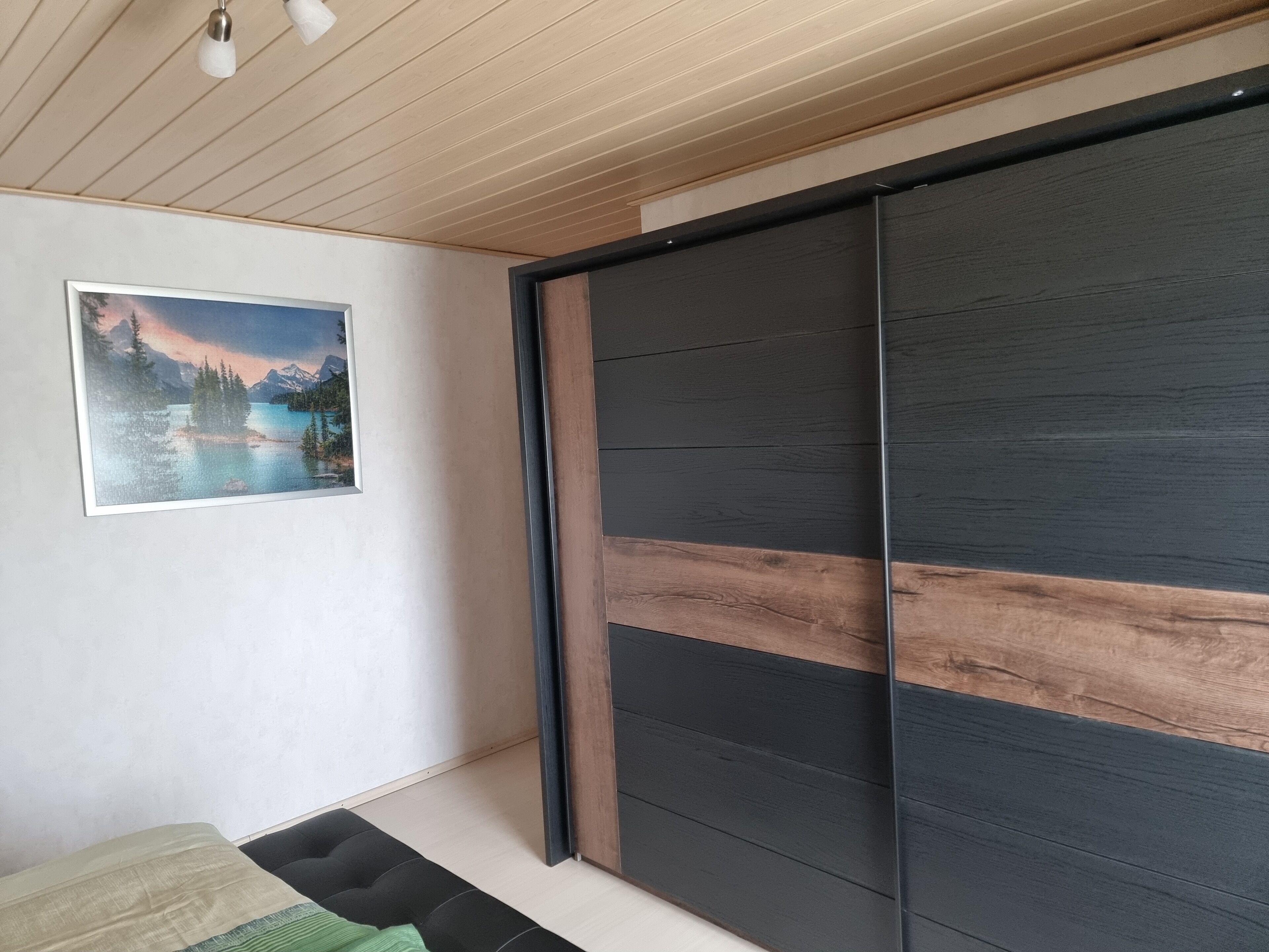 A bedroom with a bed, a sliding door, a framed picture on the wall, and a ceiling with wooden panels.