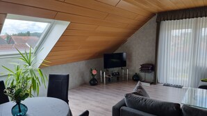 Una sala de estar moderna con techo inclinado, una mesa de comedor redonda, sofás oscuros, un televisor sobre un pedestal de vidrio y un amplio ventanal con cortinas.