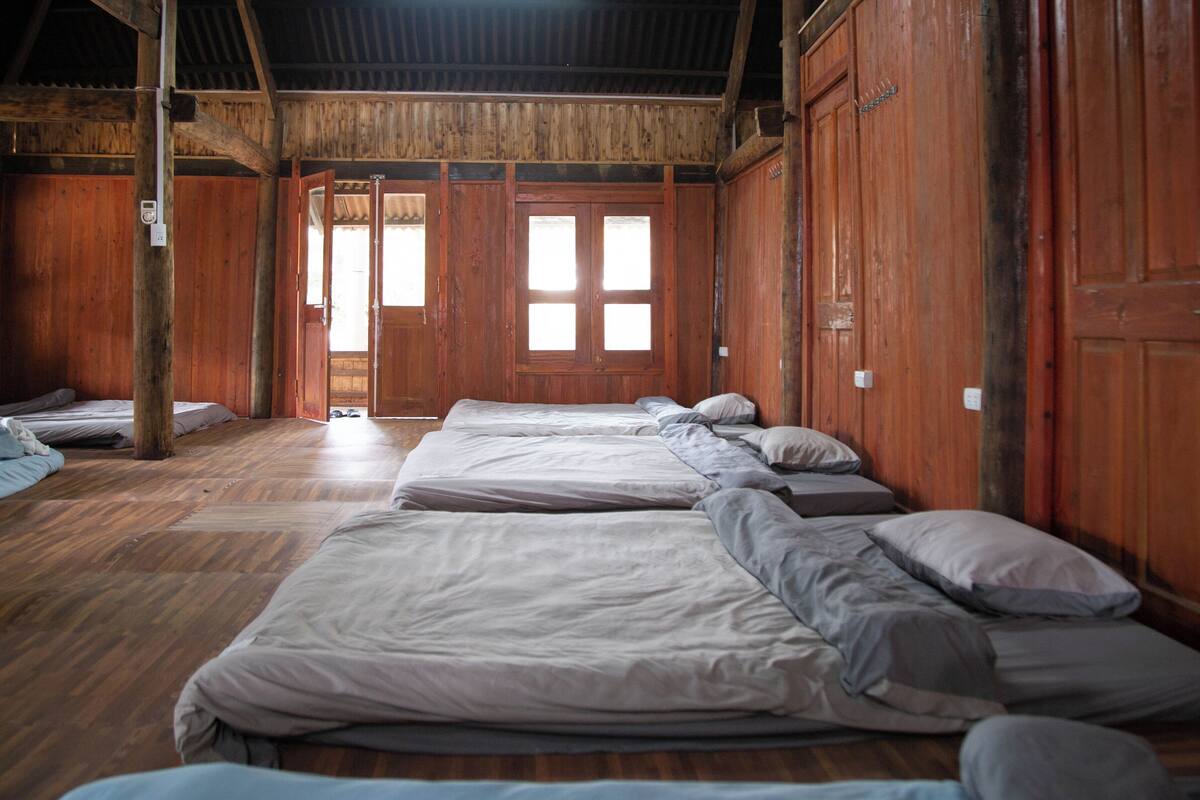 A rustic wooden room with multiple beds aligned against the walls, a wooden ceiling, and large windows allowing natural light.