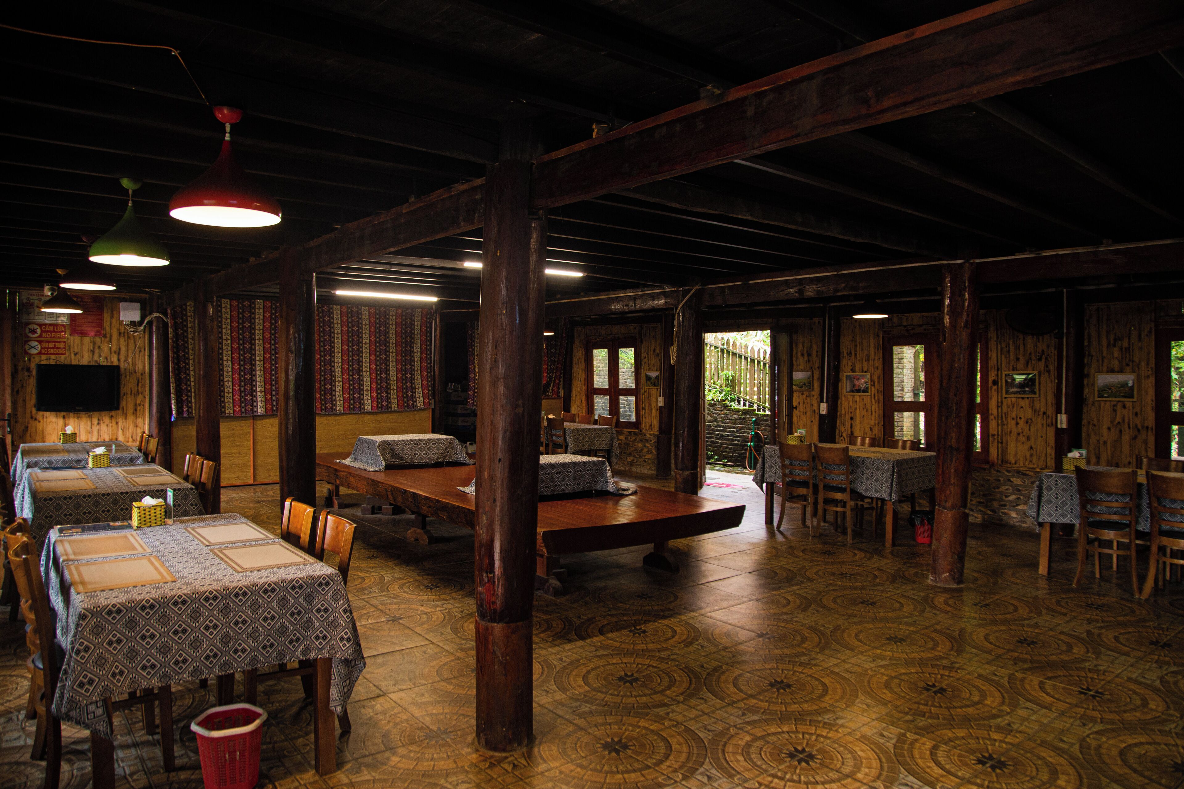 A rustic dining area with wooden tables and chairs, patterned tablecloths, and hanging pendant lights.