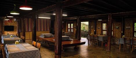 A rustic dining area with wooden tables and chairs, patterned tablecloths, and hanging pendant lights.