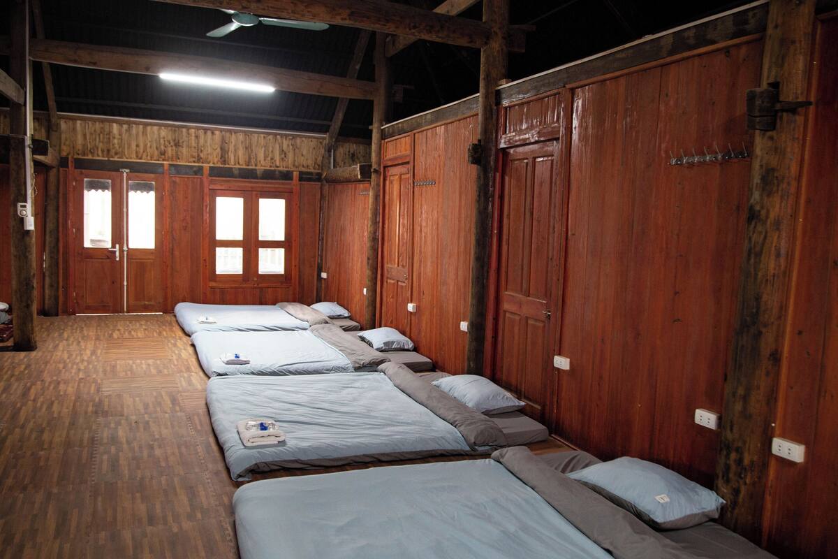 A row of wooden beds with white linens, a wooden floor, and a wooden ceiling with exposed beams.