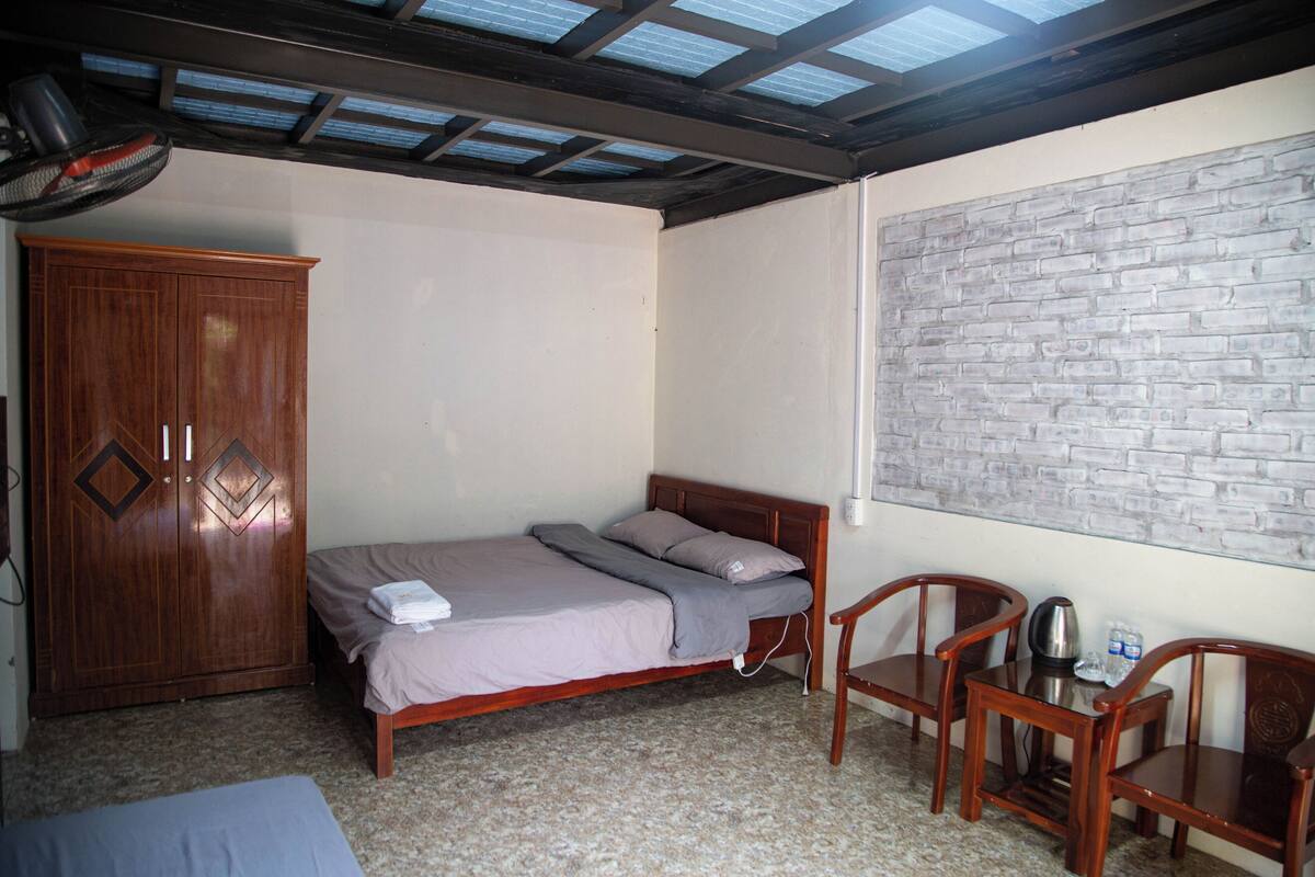 A bedroom with a bed, a wardrobe, two chairs, a table with a kettle, and a brick-patterned wall.