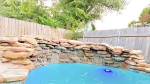 A swimming pool with a stone waterfall feature and a wooden fence in the background. - Pool House near Downtown Round Rock (Round Rock)