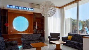 A modern living room with a large round window, two patterned sofas, a wooden coffee table, and a decorative chandelier. - Spacious & Private 6 bed villa close to Peyia - 5bed villa + 1bed granny annex (Peyia)