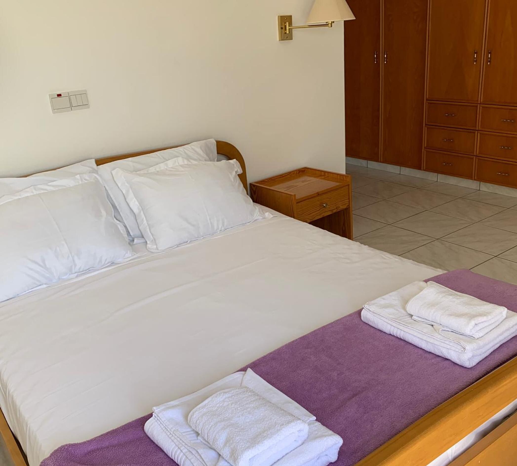 A neatly made bed with white linens, a purple towel on the side, and a wooden nightstand with a lamp.