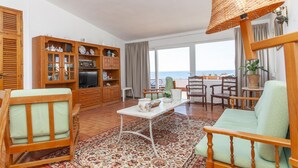 Ein Wohnzimmer mit einer Couch, Sesseln, einem Couchtisch und einem Fernseher. Ein großes Fenster bietet Blick auf das Meer.