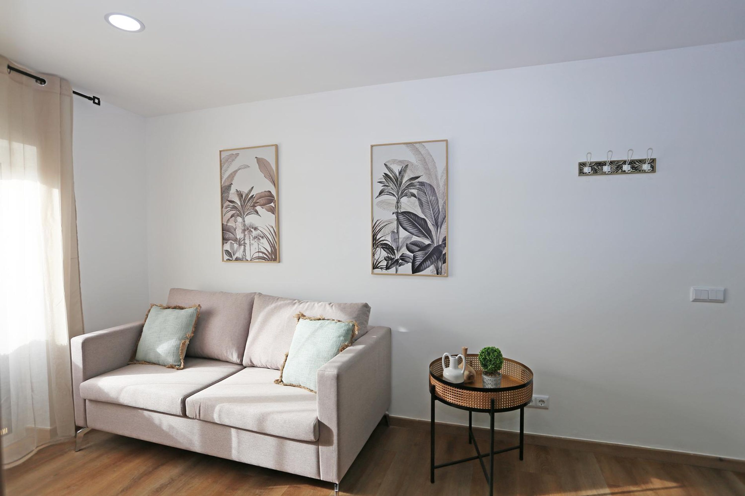 A living room with a sofa, two framed botanical prints, a small side table with a plant, and a wall-mounted light switch.