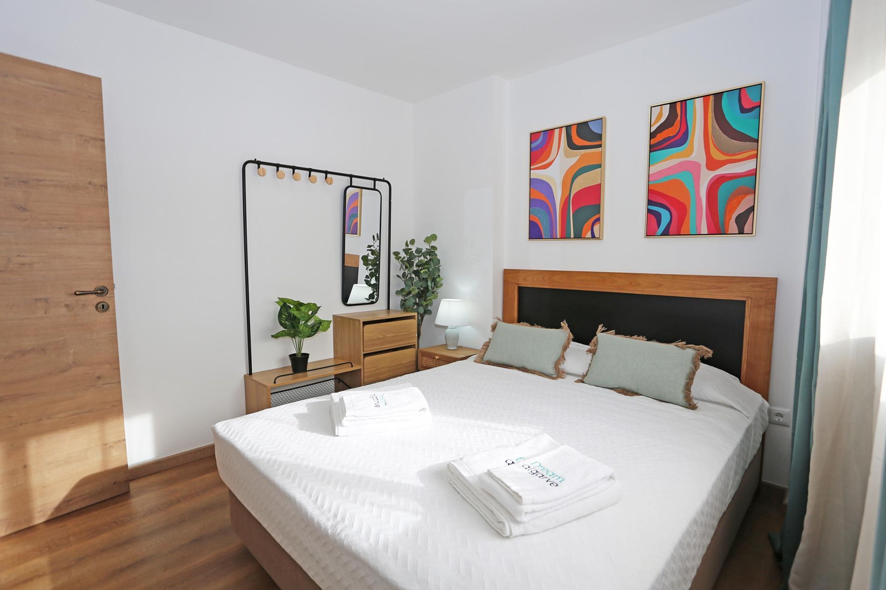 A neatly made bed with white linens and towels, a wooden headboard, a bedside table with a lamp, a mirror, and two abstract paintings on the wall.