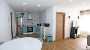 Een moderne keuken met een koelkast, magnetron en kookplaat. Er is een eettafel met stoelen en een kleine prullenbak.